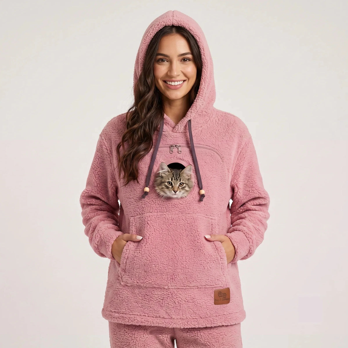 Close-up of a woman wearing the pink Dolgum Cozy Cat Hoodie with a tabby cat peeking out of the front pouch