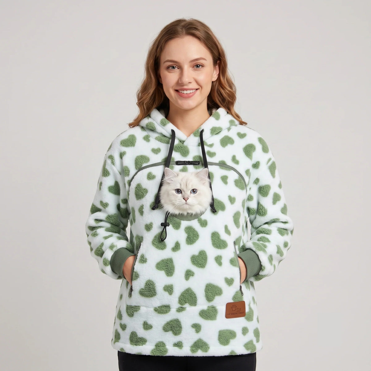 Woman wearing the green heart-print Dolgum Cozy Cat Hoodie with a white cat in the front pouch