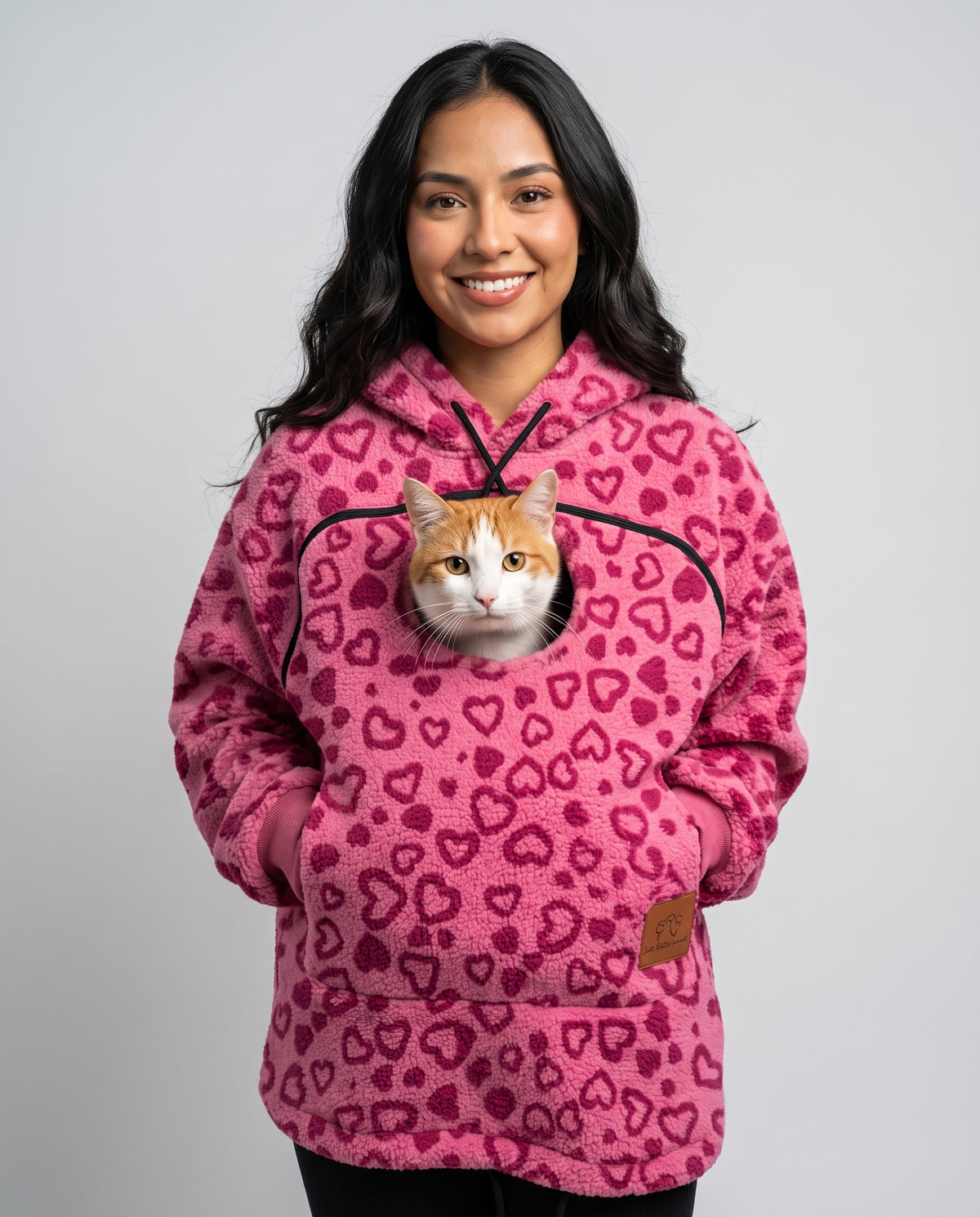 Woman wearing the pink heart-print Dolgum Cozy Cat Hoodie with an orange and white cat in the front pouch