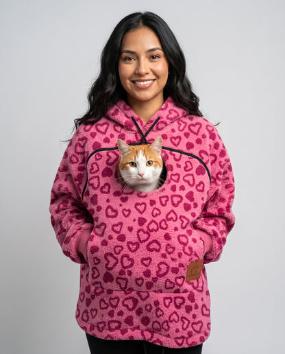 Woman wearing the pink heart-print Dolgum Cozy Cat Hoodie with an orange and white cat in the front pouch